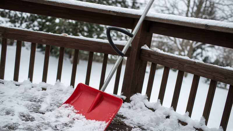 A red snow shovel with a second curved handle leaning on a snowy deck railing, in Grand Rapids, Michigan